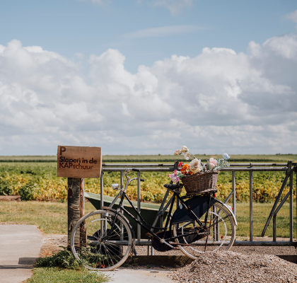 Tegen een hek met een bordje 'Slapen in de KAPschuur' staat een fiets geparkeerd, met achterop een mand vol bloemen.