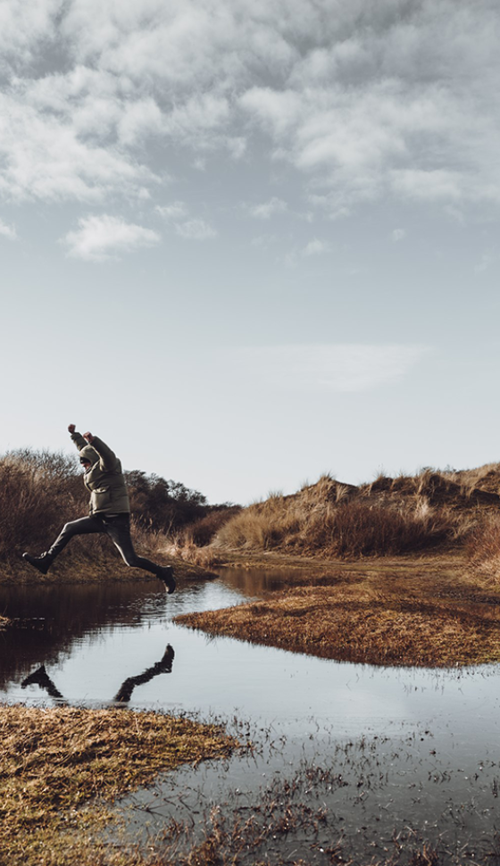 Onder een grijze, bewolkte lucht, springt een man over een grote plas heen met zijn armen in de lucht